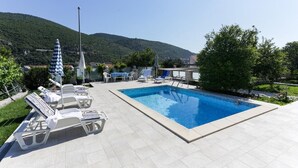 En swimmingpool med solstolar och parasoller, omgiven av grönska och en kulle i bakgrunden.