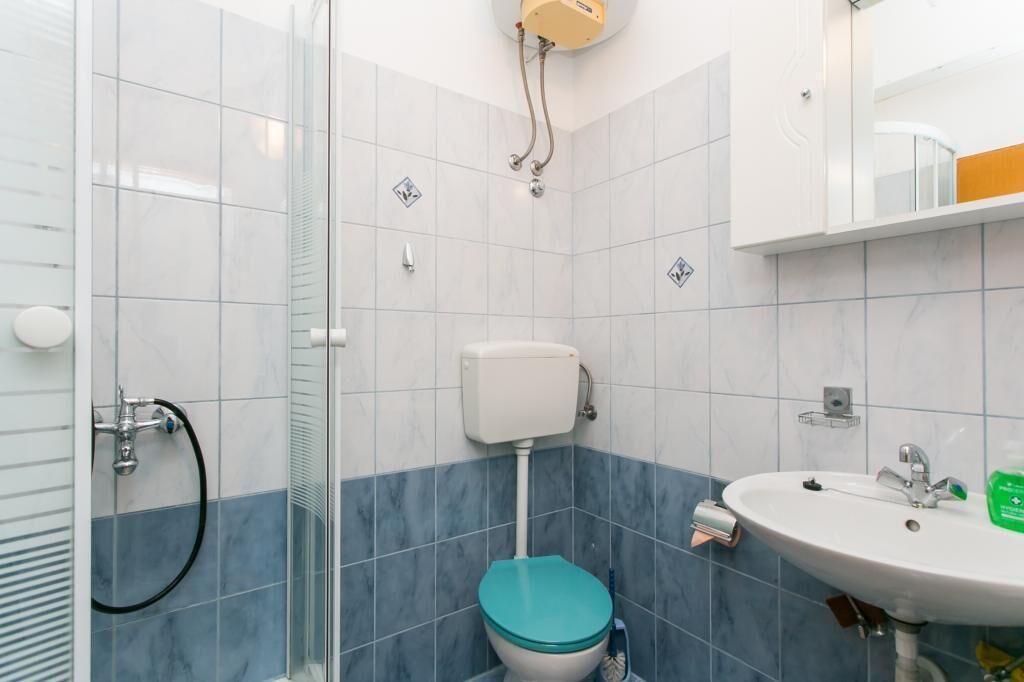 Ein Badezimmer mit Duschkabine aus Glas, Toilette mit blauem Sitz, weißem Waschbecken und Spiegel darüber.