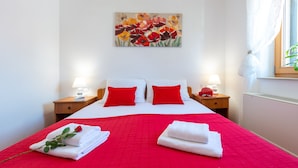 A hotel room with a large bed, red bedding, two bedside tables with lamps, a painting on the wall, and a window with a view.
