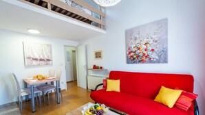A living room with a red sofa, a dining table with chairs, a fruit bowl, and a painting on the wall.