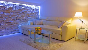 A modern living room with a stone wall, a beige sofa, a glass coffee table with fruit, and a lamp on a side table. - Big Blue Apartments - One-Bedroom Apartment with Balcony and Sea View (Big Blue 2) (Dubrovnik)