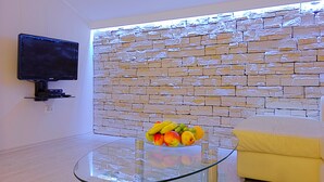 A modern living room with a stone wall, a glass coffee table with fruit, a flat-screen TV, and a yellow sofa. - Big Blue Apartments - One-Bedroom Apartment with Balcony and Sea View (Big Blue 2) (Dubrovnik)