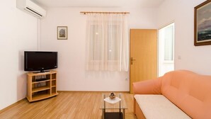 A living room with a television on a wooden stand, a sofa, a small table with a decorative item, a window with sheer curtains, and a door.