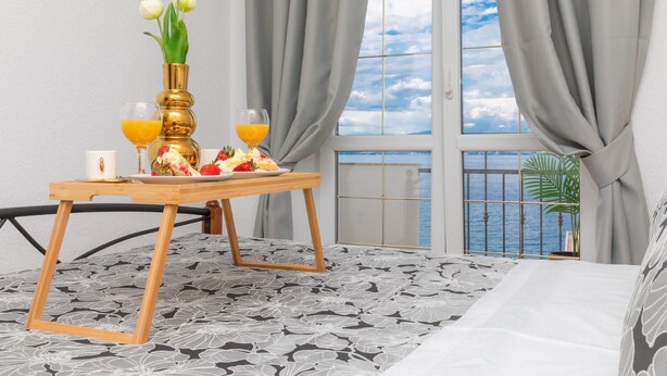 Une chambre avec un lit, une table en bois sur laquelle sont disposés des éléments pour le petit-déjeuner, un vase rempli de fleurs et une vue sur la mer, visible à travers une fenêtre ornée de rideaux.