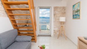 Un salon moderne avec un canapé gris, un escalier en bois, une vue sur la mer par une porte vitrée, un mur en pierre et une lampe.
