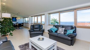 Un vaste espace de vie comprenant un canapé, une table basse et un coin repas avec une table et des chaises. De grandes fenêtres offrent une vue sur la mer.