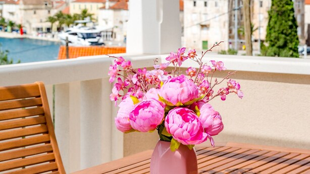 A balcony with a wooden table and a vase of pink flowers, overlooking a coastal town with red-roofed buildings and a church spire. - Apartments Sunny Days - Comfort Two Bedroom Apartment with Terrace and Sea View (A1) (ST) (Milna)