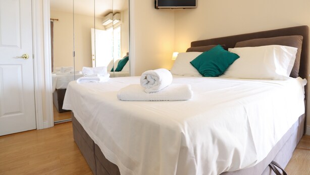 Una cama bien hecha con sábanas blancas, una toalla enrollada, un cabecero marrón, almohadas color turquesa y un televisor de pantalla plana montado en la pared.