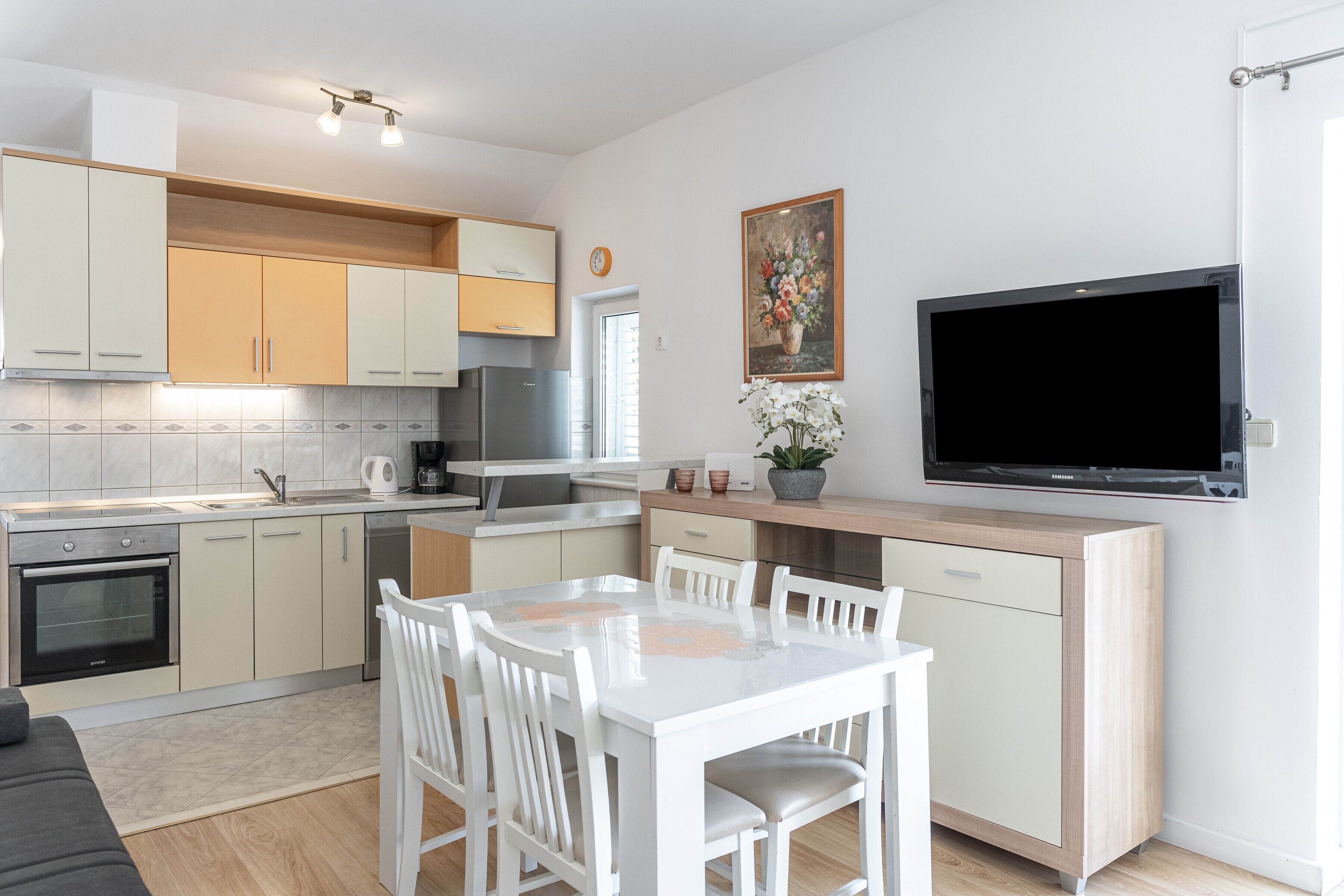 Une cuisine moderne avec des meubles blancs, un coin repas comprenant une table et des chaises blanches, un grand téléviseur à écran plat et une plante en pot.