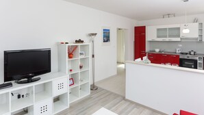 Una sala de estar moderna con un televisor de pantalla plana, estanterías blancas, una lámpara de pie y una cocina con armarios rojos y encimeras blancas.