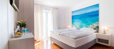 A modern bedroom with a large bed, a bedside table with a lamp, a painting of a beach, and a window with curtains.