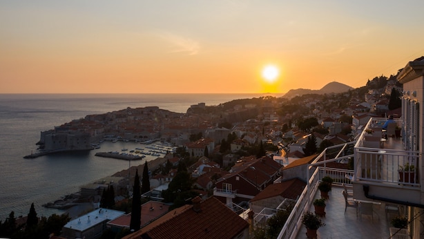 Une ville côtière abritant un château imposant au bord de la mer, un balcon orné de plantes en pot et offrant une vue dégagée sur le coucher de soleil sur l’eau.