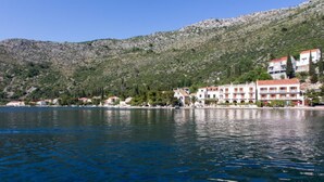 Un pueblo costero con edificios a lo largo del paseo marítimo, un mar azul cristalino y un telón de fondo montañoso.