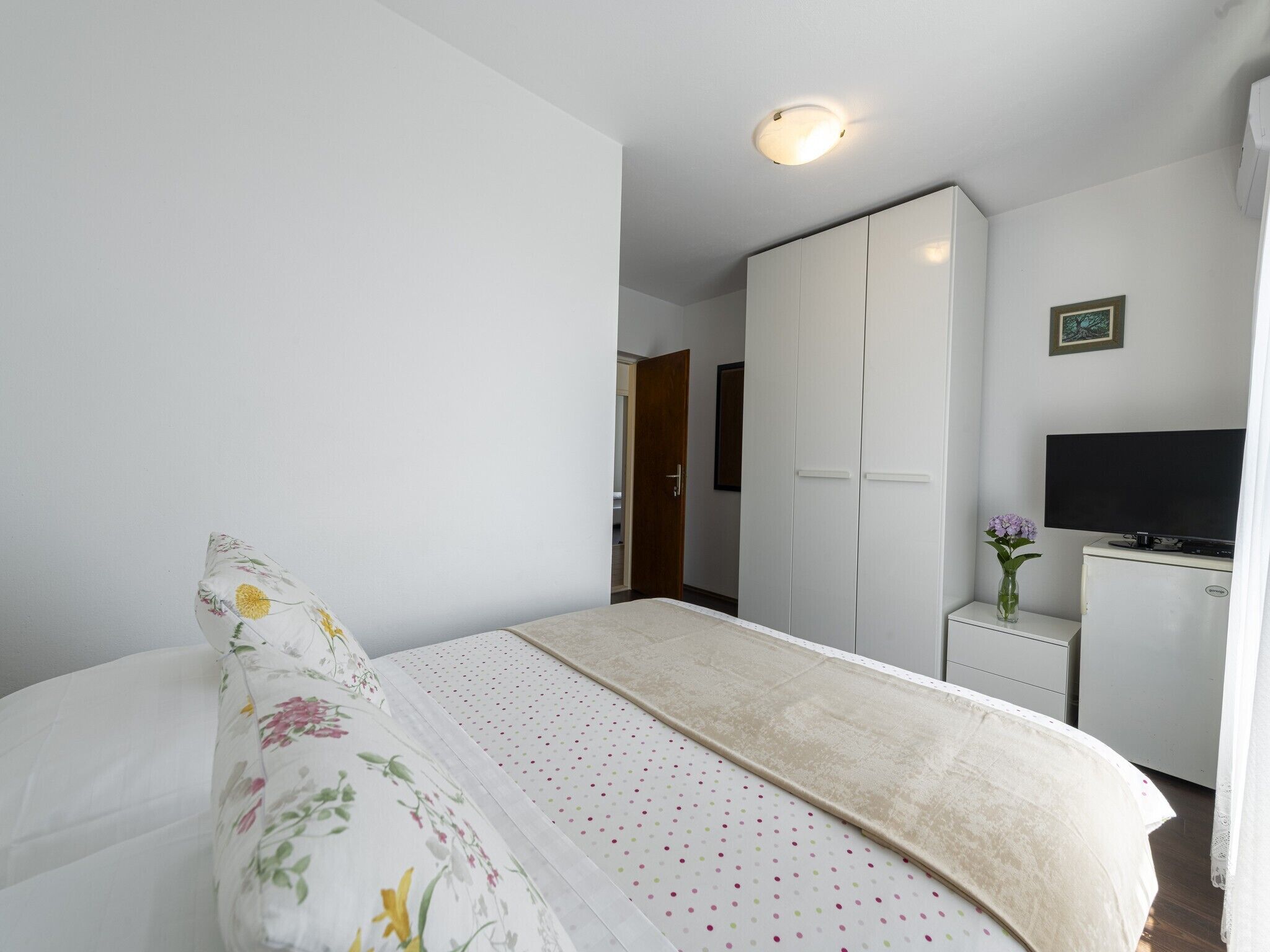 Une chambre à coucher avec un lit, une armoire, un bureau avec une télévision et un petit vase avec des fleurs.