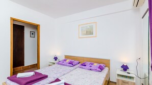 Ein ordentlich bezogenes Bett mit violett-weißer Bettwäsche, ein kleiner Nachttisch mit Lampe und eine Holztür.