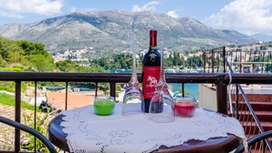 Ein Tisch mit einer Weinflasche, zwei Gläsern und zwei Getränken auf einem Balkon mit Blick auf eine gebirgige Landschaft.