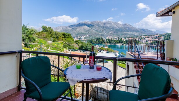 Ein Balkon mit zwei grünen Stühlen, einem kleinen Tisch mit Spitze und einer Weinflasche. Im Hintergrund ist ein See, Berge und eine Stadt zu sehen.