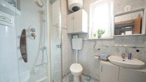 Une salle de bain avec une douche à parois de verre, des toilettes blanches, un lavabo avec un miroir au-dessus et une fenêtre donnant sur un espace verdoyant.