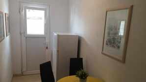 Un petit coin repas avec une table jaune, deux chaises noires, un réfrigérateur et une porte qui donne sur une pièce plus lumineuse.