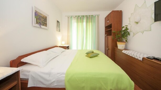 Une chambre à coucher comprenant un lit, une table de chevet, une lampe, une fenêtre avec des rideaux, un téléviseur fixé au mur, une armoire en bois et une plante en pot.