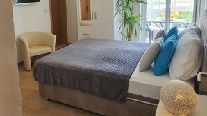 Ein Schlafzimmer mit Bett, Fernseher, Kleiderschrank, Stuhl, Pflanze und Blick ins Freie.
