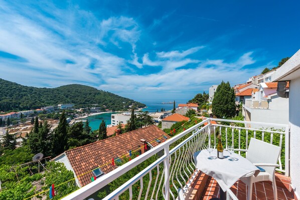 A balcony with a table and chairs, overlooking a coastal town with terracotta roofs and a clear blue sky.