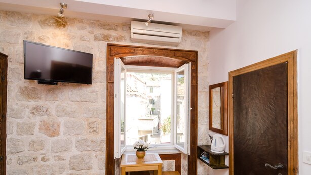 Habitación de paredes de piedra con una televisión de pantalla plana montada, una mesa de madera con un banco, una ventana con vistas a edificios y una puerta con espejo.