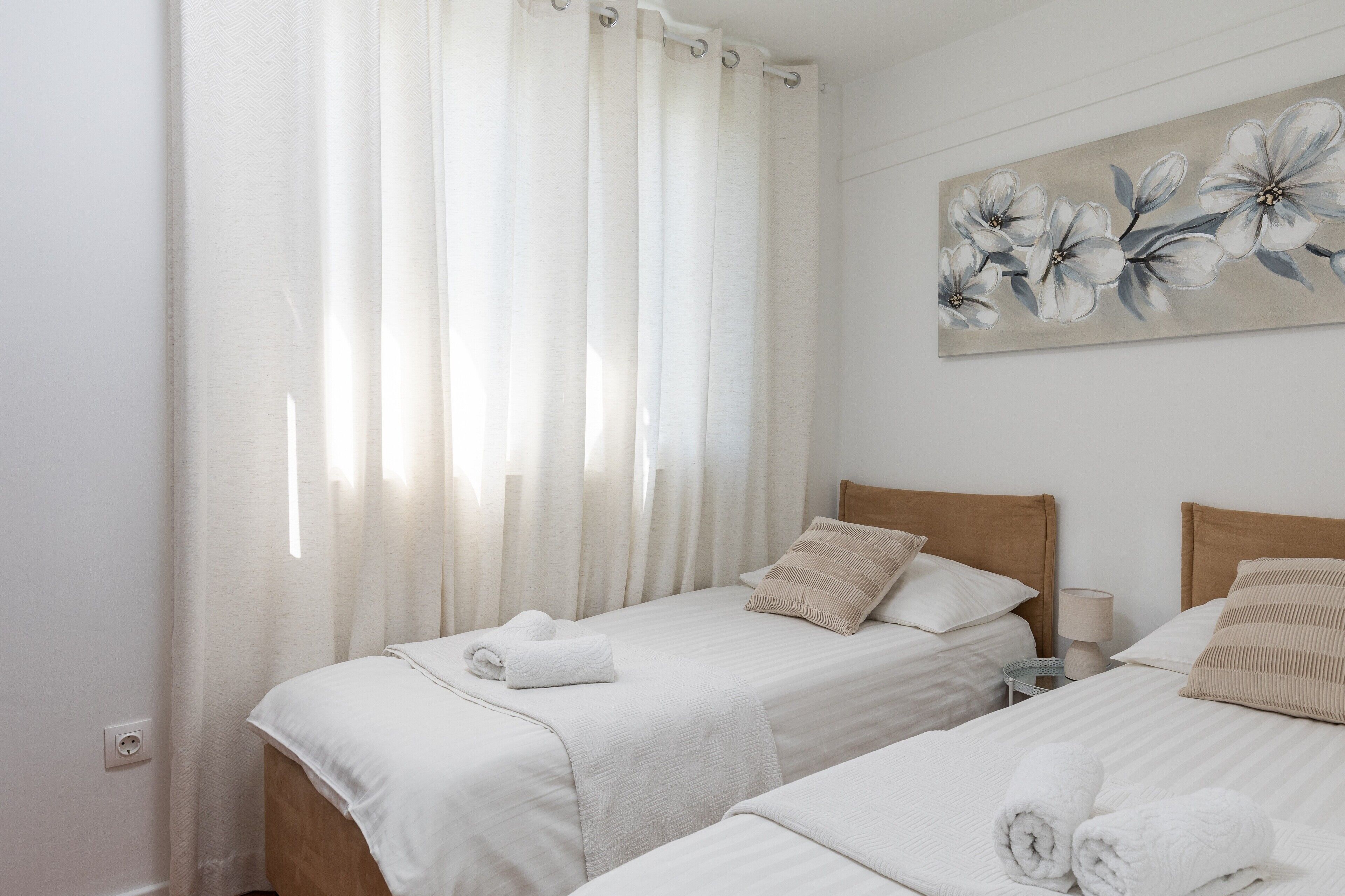 A hotel room with two beds, white bedding, a painting of flowers on the wall, and a window with sheer curtains.