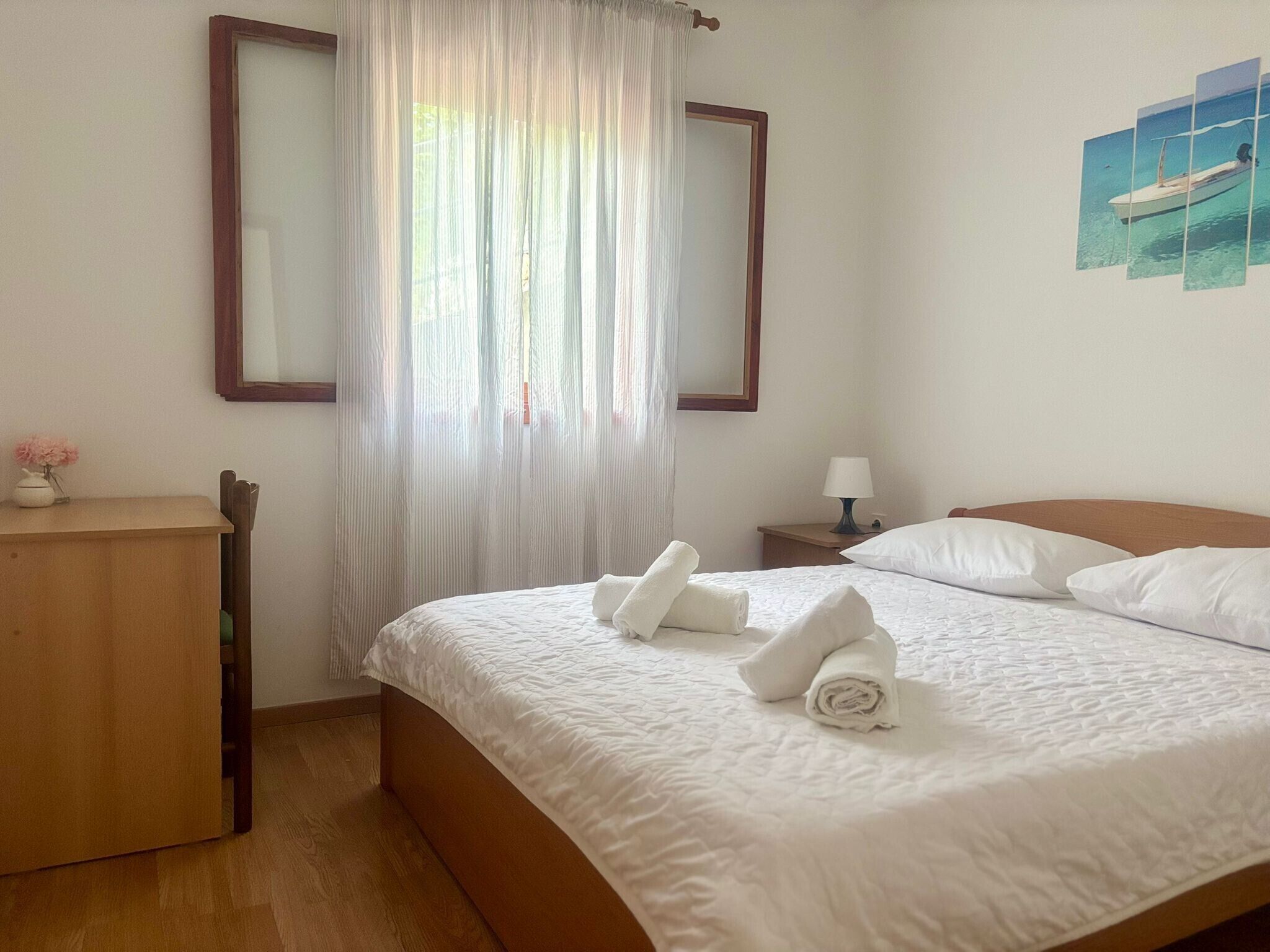 A neatly made bed with white linens and towels, a wooden nightstand with a lamp, a wooden desk with a vase of flowers, and a large window with sheer curtains.