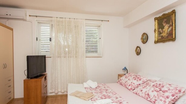 Ein Schlafzimmer mit einem Bett, einem Fernseher auf einem Standfuß, einem Fenster mit Jalousien und einem an der Wand hängenden Bild.