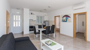 Un appartement moderne comprenant un salon, un coin repas et une cuisine. Le salon est équipé d’un canapé, d’une table à manger et de chaises. La cuisine est dotée de meubles blancs et d’une fenêtre.