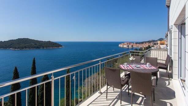 Un balcon doté de meubles en osier et d’une table, offrant une vue sur la côte, avec une île et la mer en toile de fond.