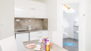 Eine kompakte Küche mit weißen Schränken, einer grauen Spritzschutzleiste und einem kleinen Spülbecken. Ein Essbereich mit Tisch, Stühlen und einer Obstschale ist vorhanden. Ein Badezimmer ist sichtbar, mit Toilette und Duschbereich.