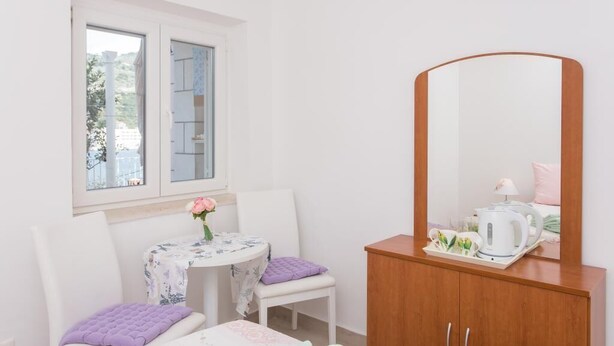 Une petite pièce propre, avec une fenêtre, une table recouverte d’une nappe à motifs floraux, une chaise, un meuble en bois et un miroir.