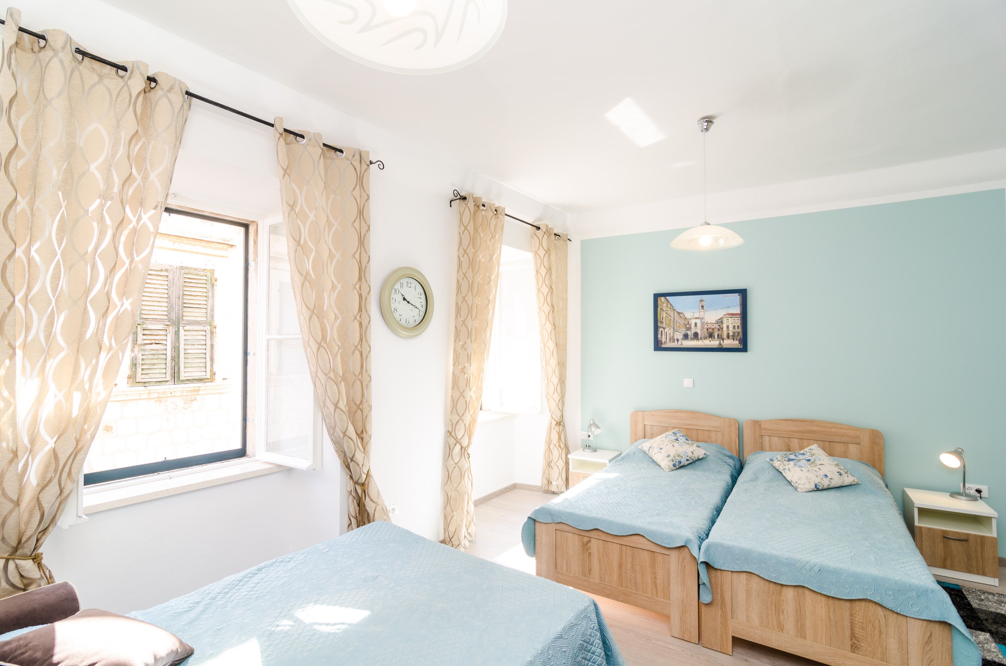 Un dormitorio con dos camas, una ventana con cortinas, un reloj, una mesita de noche con lámpara y un cuadro en la pared.