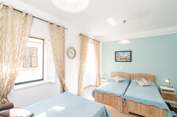 Une chambre avec deux lits, une fenêtre avec des rideaux, une horloge, une table de chevet avec une lampe et un tableau encadré au mur.