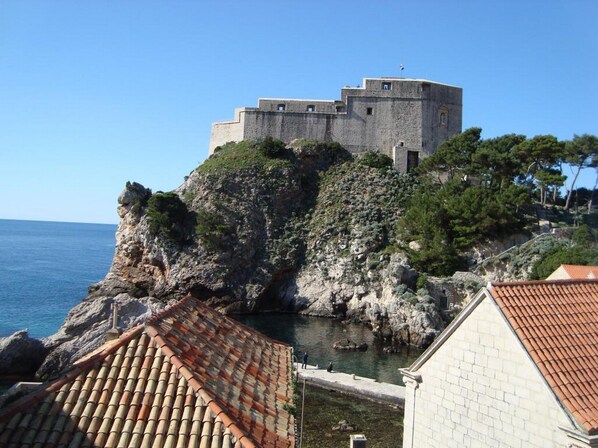 Une forteresse historique en pierre, perchée sur une falaise surplombant la mer, avec un petit port et des toits en tuiles traditionnelles à proximité.