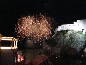 Une scène nocturne avec des feux d’artifice illuminant un paysage rocheux et un bâtiment blanc doté d’un toit plat.