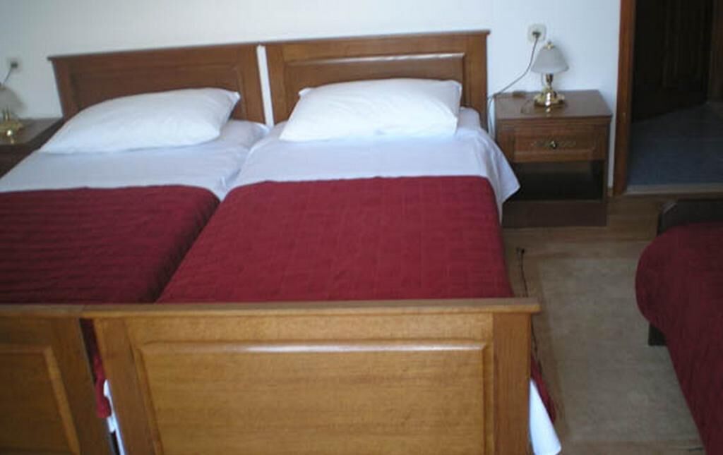 Ein Doppelbett mit Holzrahmen und roter Bettwäsche. Ein Nachttisch mit Lampe und Schublade sind ebenfalls vorhanden.
