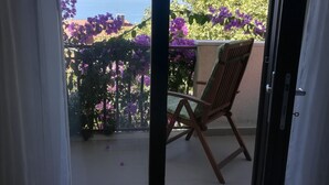 Une pièce avec un plancher en bois, une chaise et une vue sur un balcon orné de fleurs violettes et donnant sur une étendue d’eau.