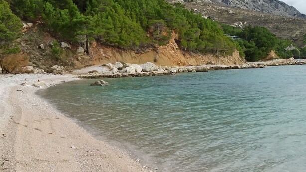 Une plage à l’eau turquoise et limpide, avec un rivage de galets et une falaise rocheuse parsemée de végétation verdoyante.