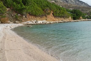 A beach with clear turquoise water, pebbly shore, and a rocky cliff with green vegetation. - Apartment Tamara- Three Bedroom Apartment with Balcony (Lokva Rogoznica)