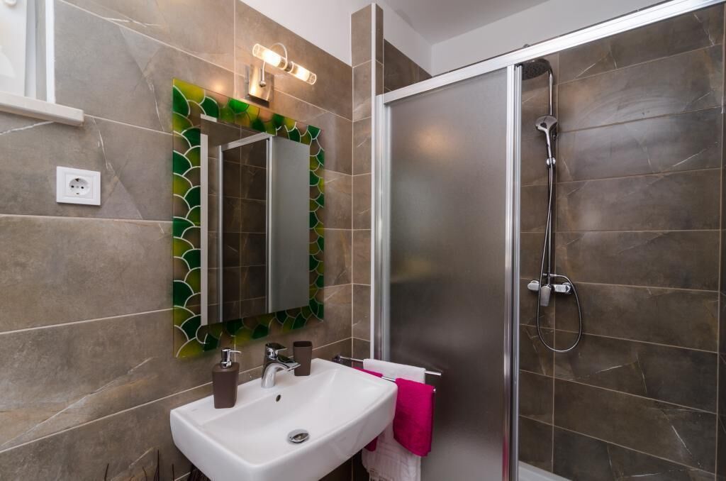 A modern bathroom with a white sink, a mirror with a green leaf pattern, and a shower with dark tiles.