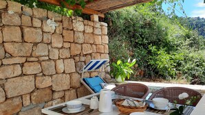 Un muro de piedra con una mesa dispuesta para comer, una silla de mimbre y una planta en maceta.