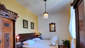 Una habitación con piso de madera, una cama con cabecera, una mesita de noche con lámpara y un cuadro colgado en la pared.