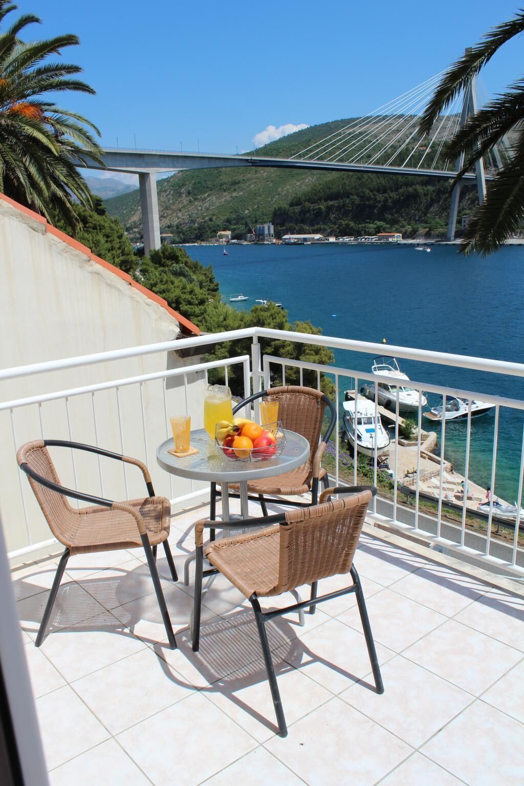 A balcony with wicker chairs and a table set with fruit and a drink, overlooking a marina and a bridge.