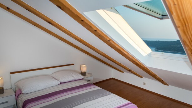 A bedroom with a sloped ceiling, a bed with a striped blanket, bedside lamps, and a view through a skylight. - Rooms Raic - Studio Apartment (Olive) (Dubrovnik)