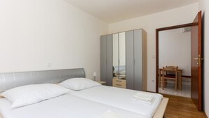 Ein Doppelbett mit weißer Bettwäsche, ein grauer Kleiderschrank und eine Holztür zu einer kleinen Sitzecke mit Tisch und Stuhl.