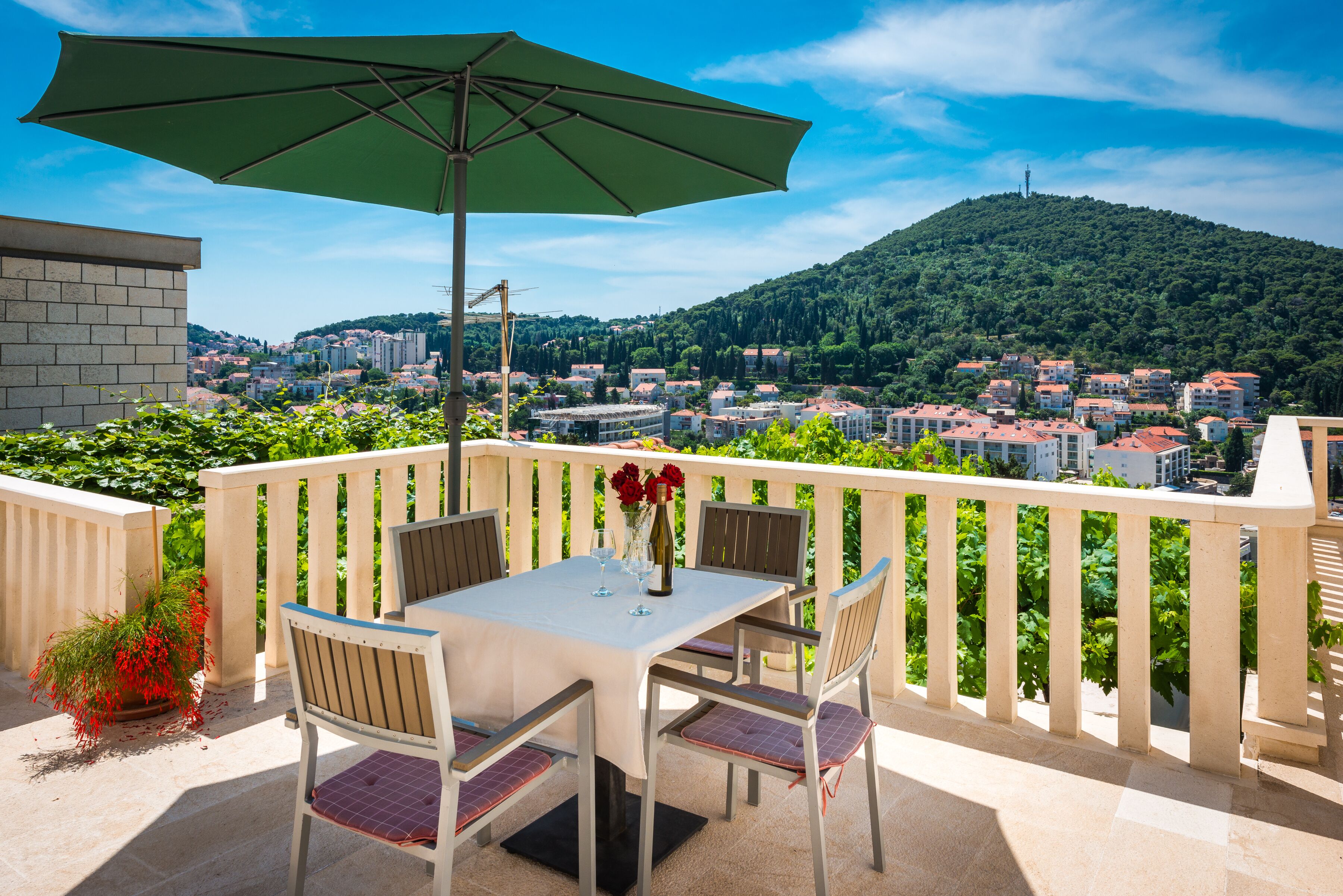 A balcony with a table and chairs, a green umbrella, and a view of a town and hills.