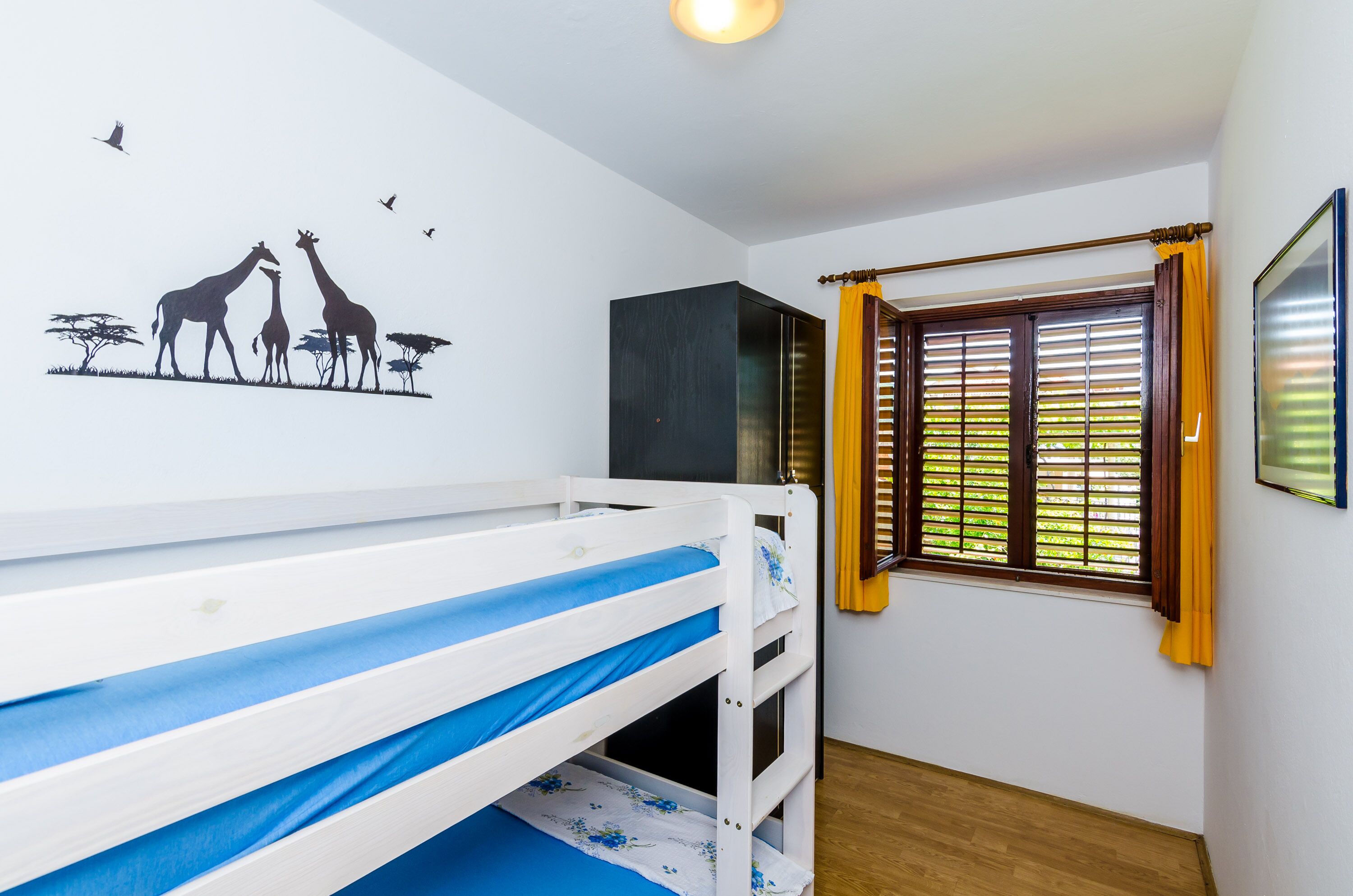 A room with a bunk bed, a window with blinds, a wall decal of giraffes, and a framed picture on the wall.